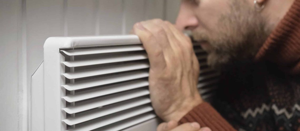 Young frozen man wearing a sweater shaking and freezing for winter cold with shock expression on face. Boy is warming up hands with breath over electric heater. Discomfort spending time at home. Heating problems. Central Heating Crises. Rise in price of Energy. Global Energy Shortage. International gas supply crisis. World crisis, export, import. Gas problems, sanctions. Increase prices