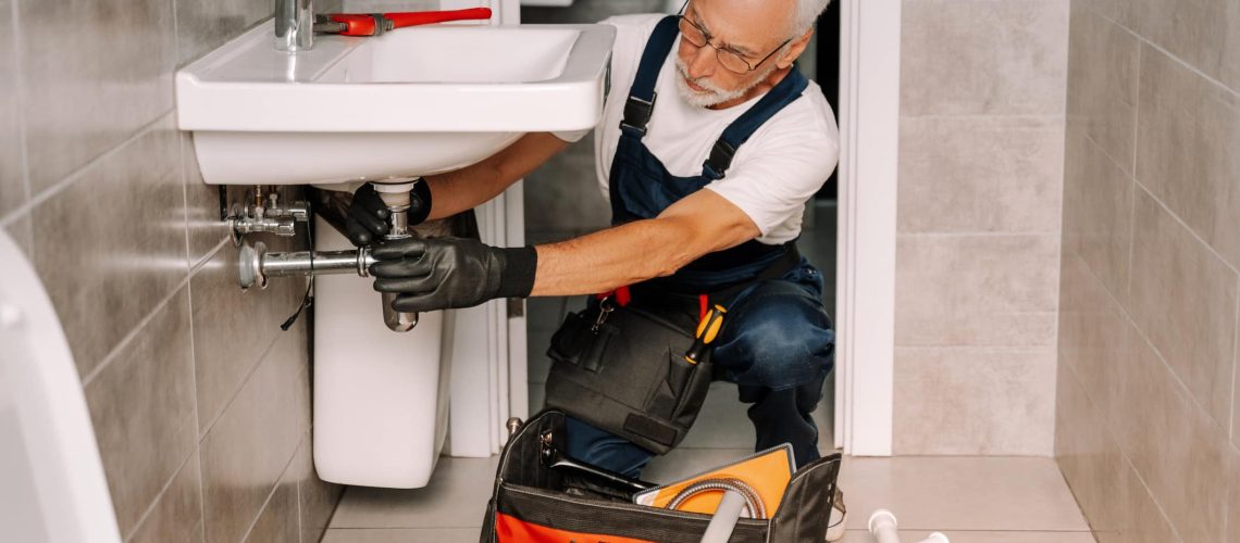 Plumber is working under a sink, installing the piping with a wrench, in a bathroom. His toolbox is open on the floor next to him