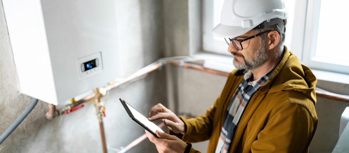 Experienced technician wearing safety gear using a tablet to evaluate a gas heating unit in a bright, industrial interior. The image showcases professional skill and modern technology in heating system maintenance.